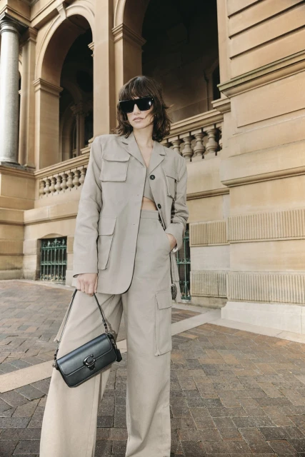 A woman in a tan blazer and wide-leg pants poses outdoors in front of a building with arches, carrying a black Coach purse.