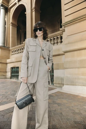 A woman in a tan blazer and wide-leg pants poses outdoors in front of a building with arches, carrying a black Coach purse.