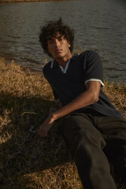 Young man with curly hair poses by a tranquil lake in a navy polo shirt and dark pants, bathed in warm light with grassy foreground.