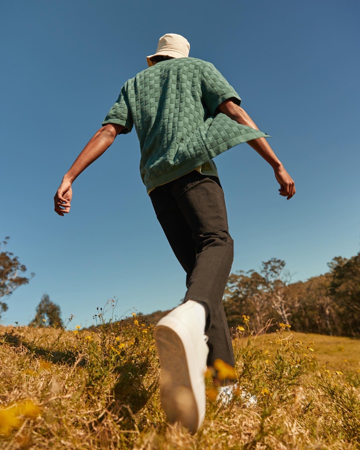 Low-angle shot of an adult man walking in a field wearing a bucket hat, green short-sleeved shirt, and black pants against a blue sky backdrop.