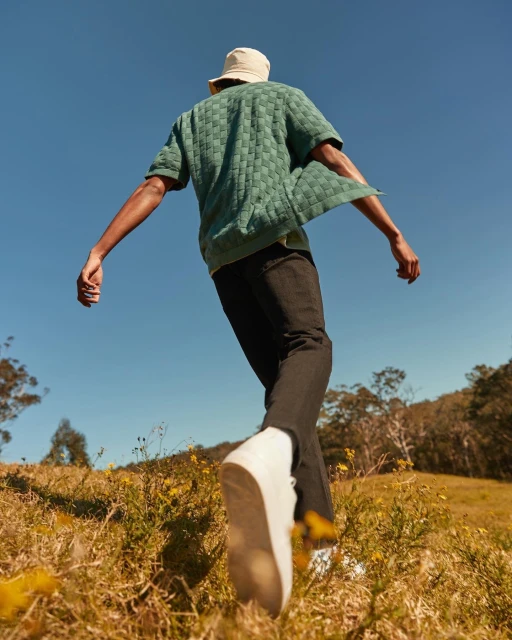 Low-angle shot of an adult man walking in a field wearing a bucket hat, green short-sleeved shirt, and black pants against a blue sky backdrop.