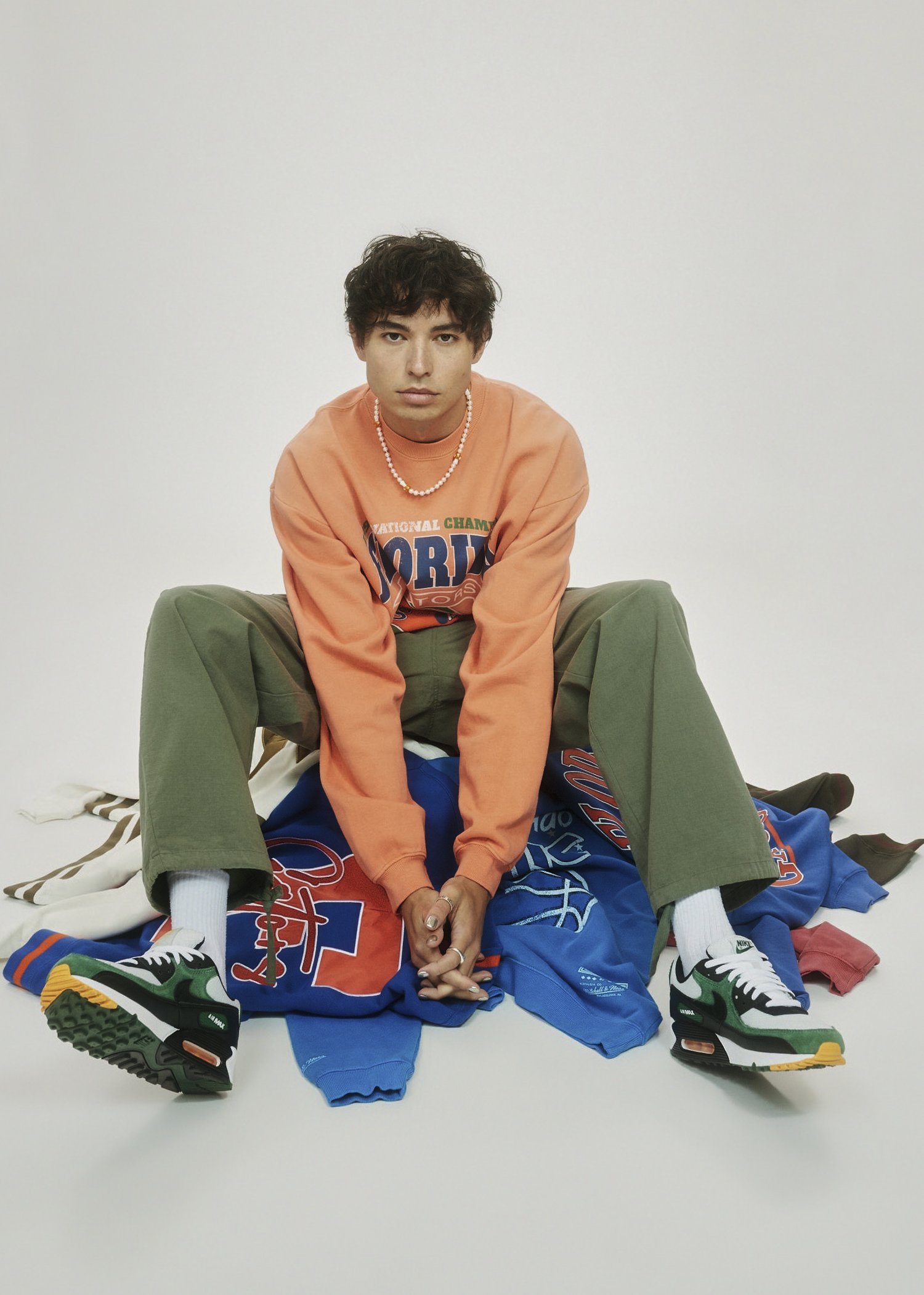 Young adult in an orange sweatshirt and green pants sits on a pile of sweatshirts and wears sneakers against a neutral background.