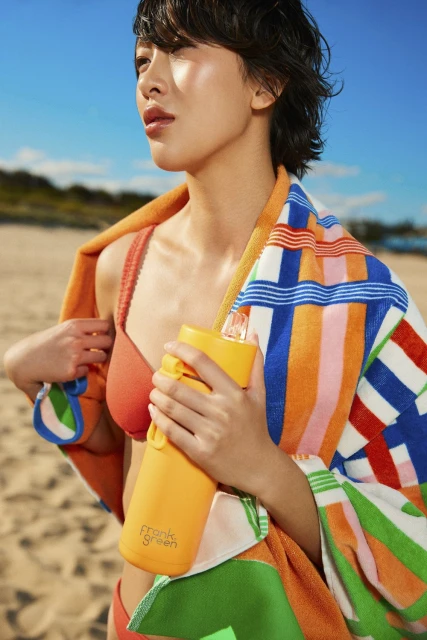 Young woman in bikini holds orange water bottle on a beach, with a striped towel around her shoulders against a sunny blue sky.