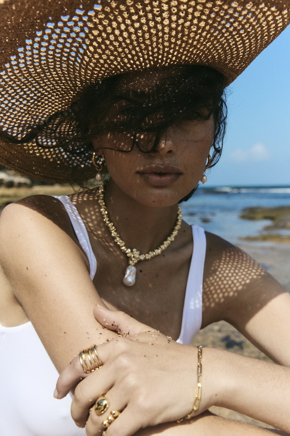 Woman wearing sun hat and jewelry on sunny beach. She has on gold earrings, necklace, and rings. Background of ocean and blue sky.