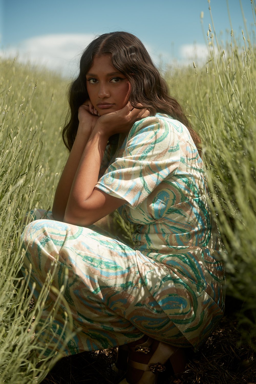 Young woman in a patterned jumpsuit poses while sitting in a grassy field with a blue sky in the background, creating a summery vibe.