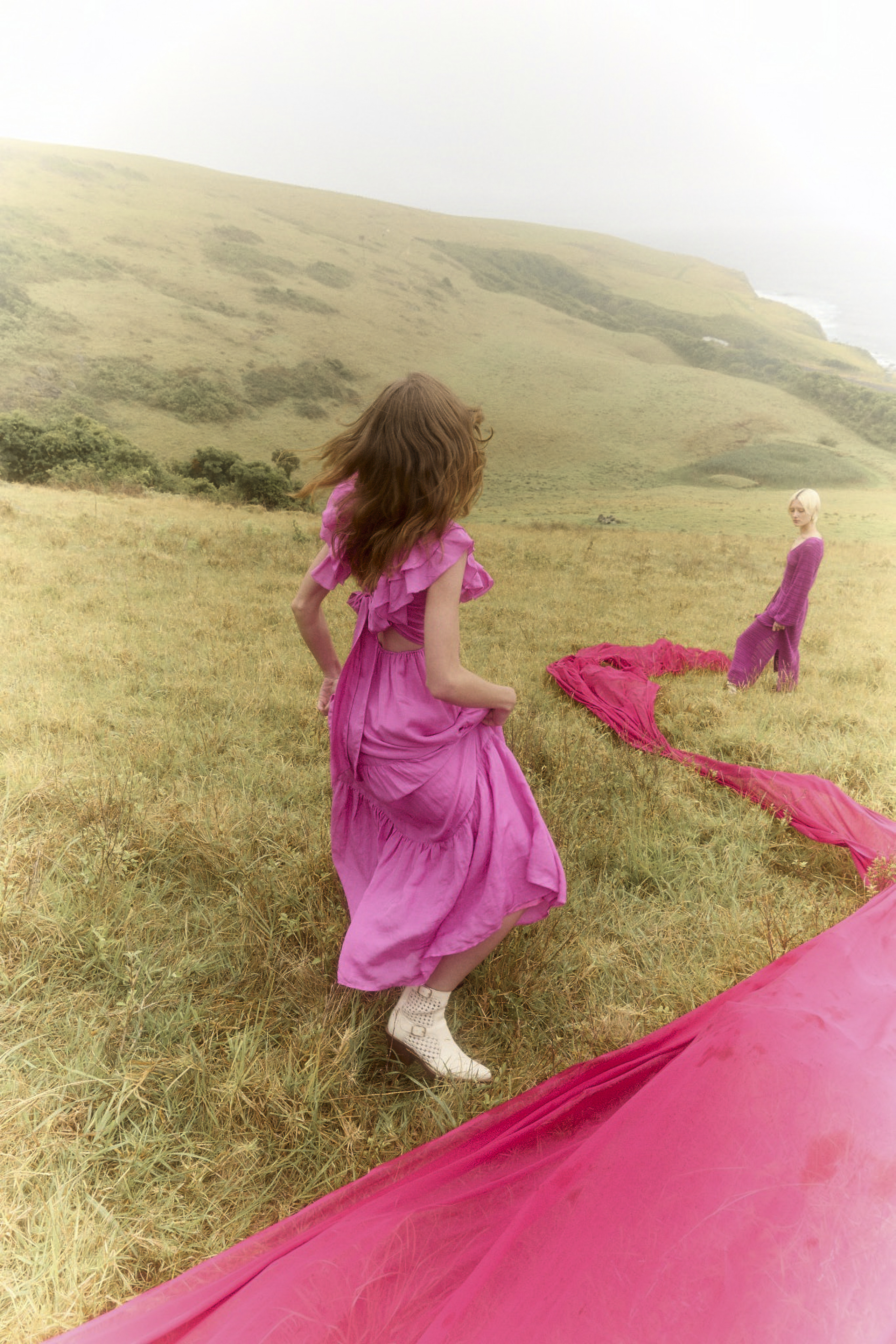Two women in pink dresses in a grassy field. One woman is facing away from the camera with a long pink cloth on the ground.