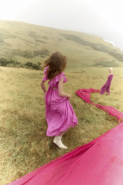 Two women in pink dresses in a grassy field. One woman is facing away from the camera with a long pink cloth on the ground.