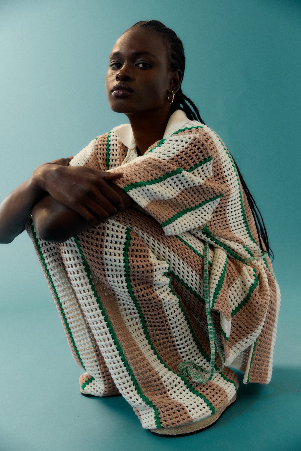 Dark-skinned woman in a crochet dress sits in a studio with turquoise background, showcasing fashion and design elements in a medium shot.