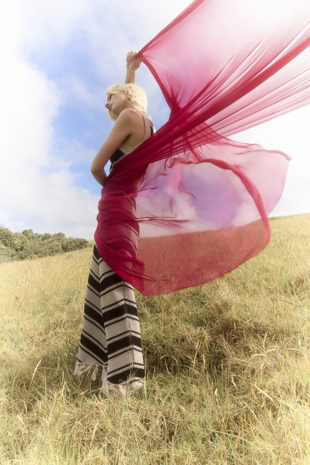 A woman with blonde hair holds a red translucent fabric on a grassy hill under a partly cloudy sky, wearing striped pants and a tank top.