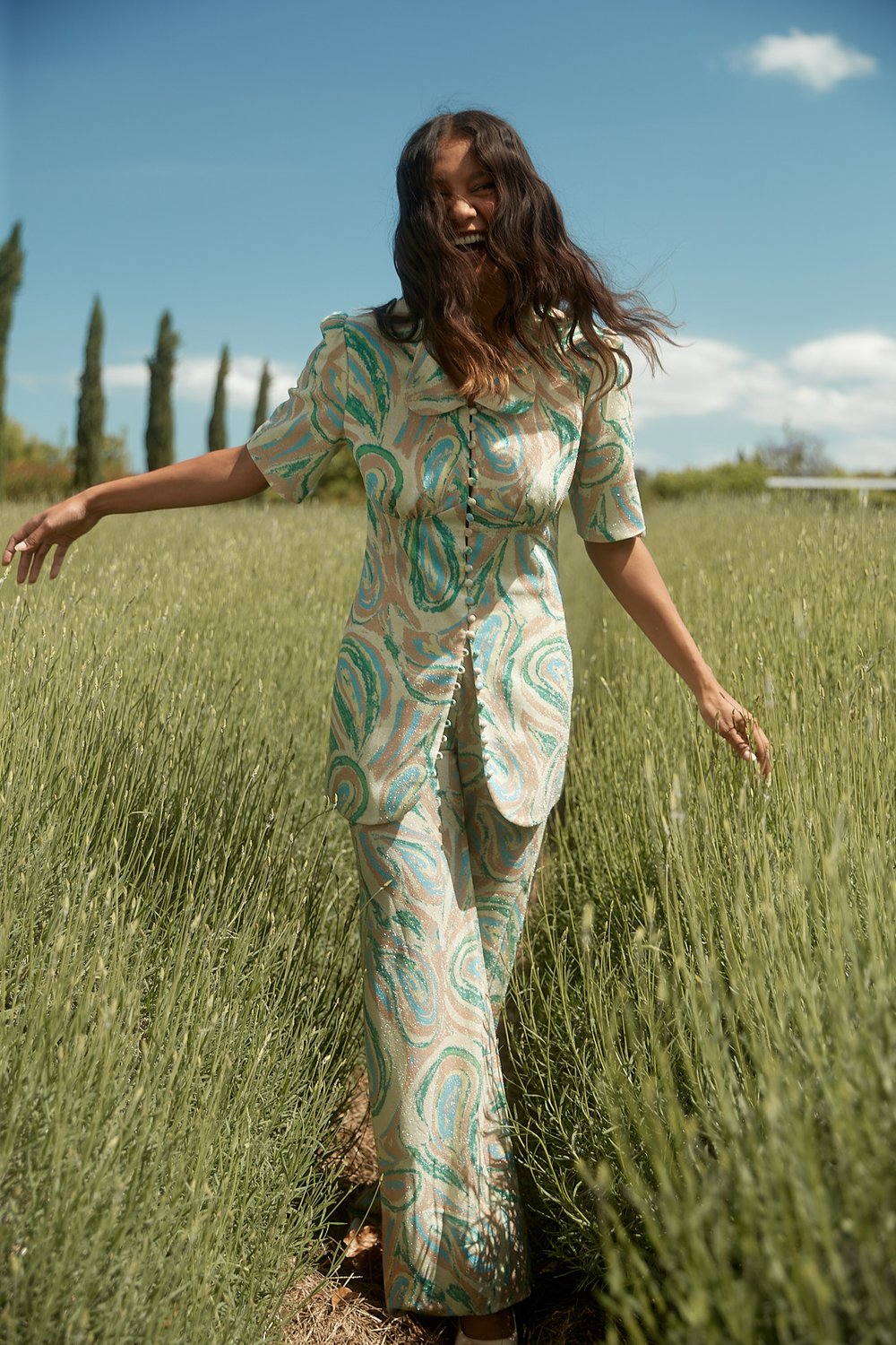 A young woman in a patterned pantsuit joyfully walks through a tall grassy field, under a sunny sky with scattered trees in the background.