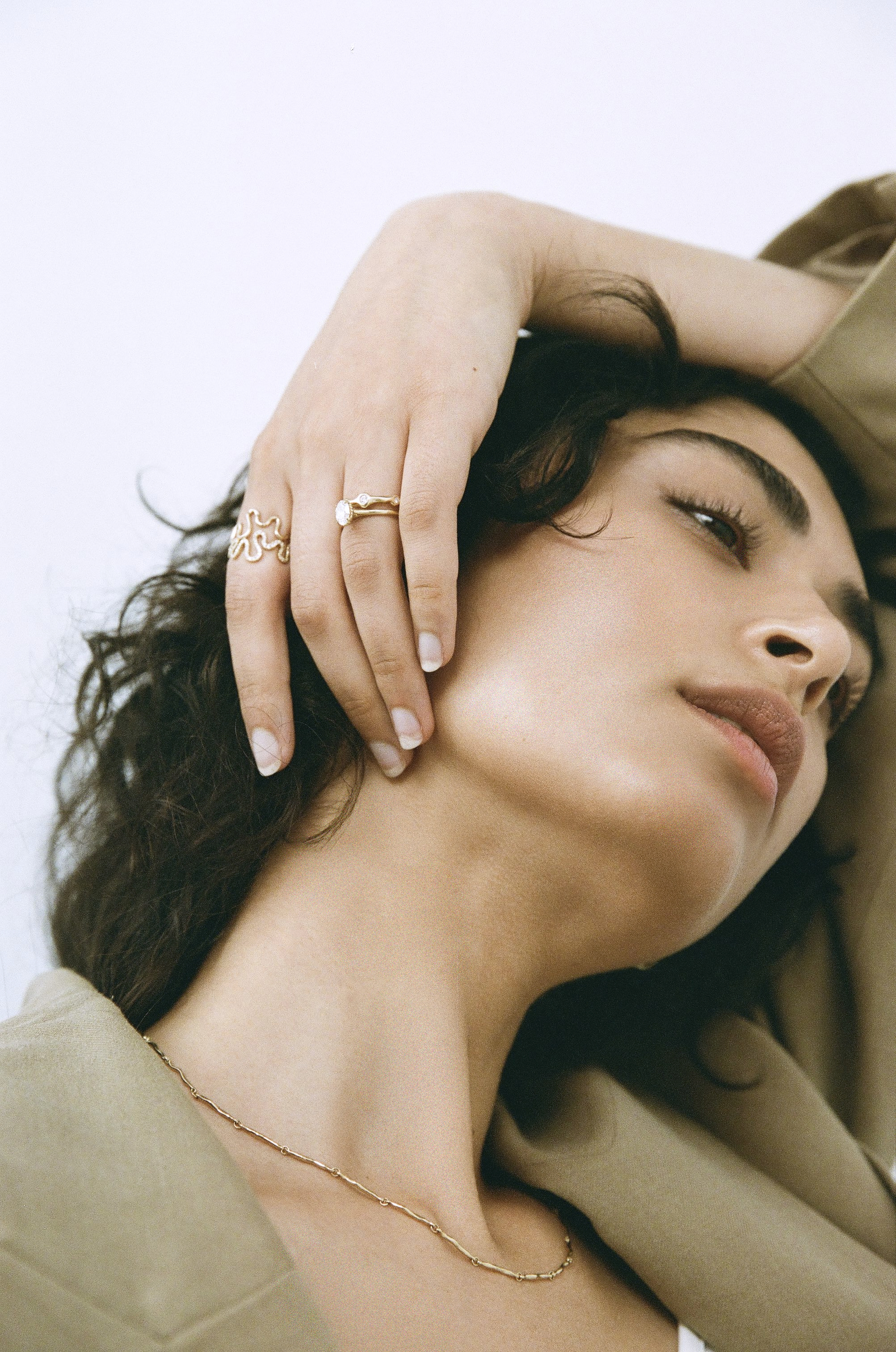 Woman with jewelry, wearing a blazer, poses in a close-up studio shot with white background. Rings and a delicate necklace are visible.