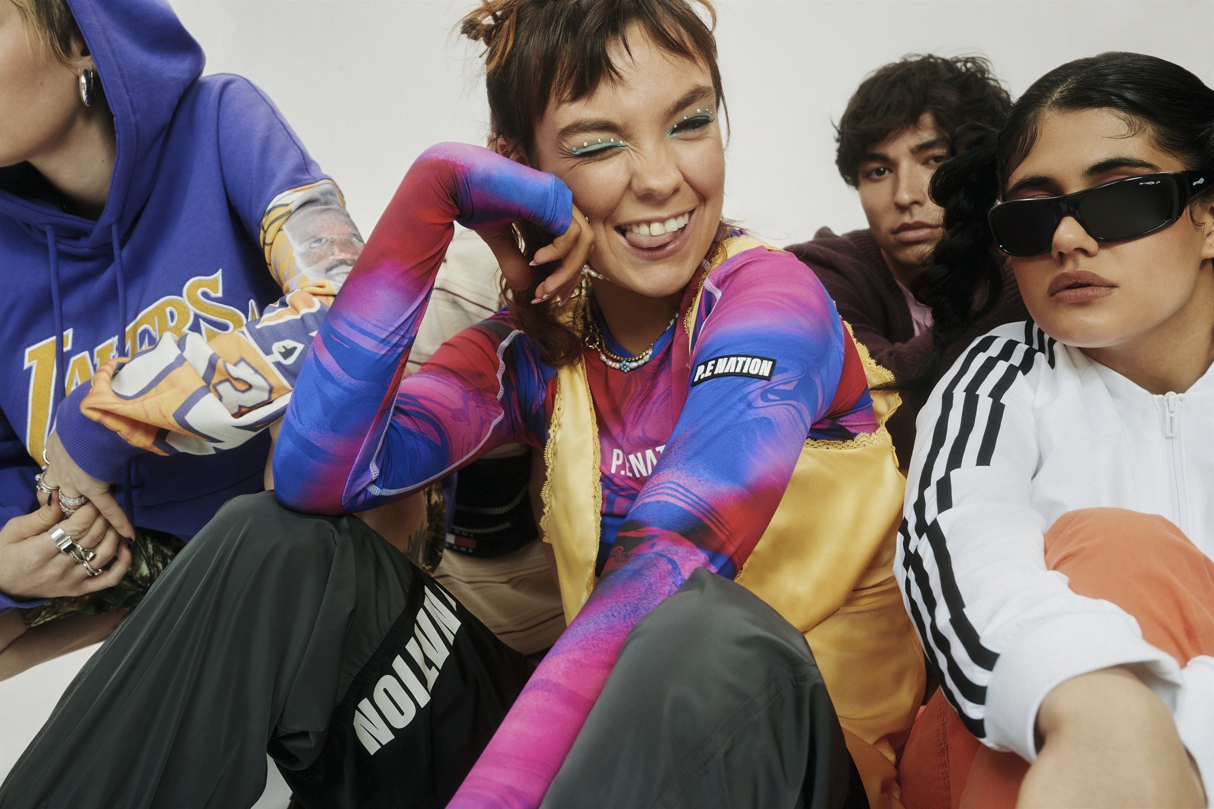 Group of five young adults indoors, wearing colorful casual clothing. One woman in the foreground is sticking her tongue out.