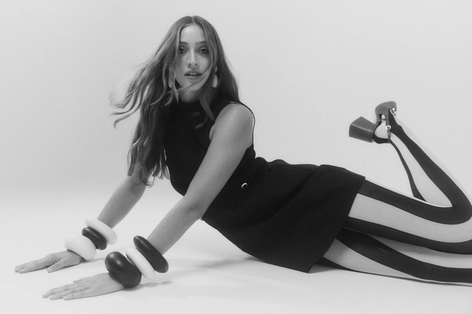Black and white portrait of a woman in a black dress and striped tights lying on a white surface with geometric bracelets.