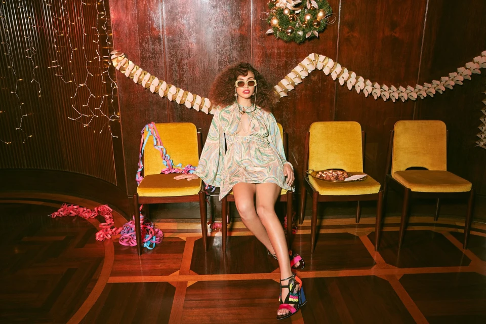 A woman with curly hair sits on a yellow chair wearing a patterned dress and sandals, with three other chairs behind her.
