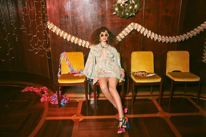 A woman with curly hair sits on a yellow chair wearing a patterned dress and sandals, with three other chairs behind her.