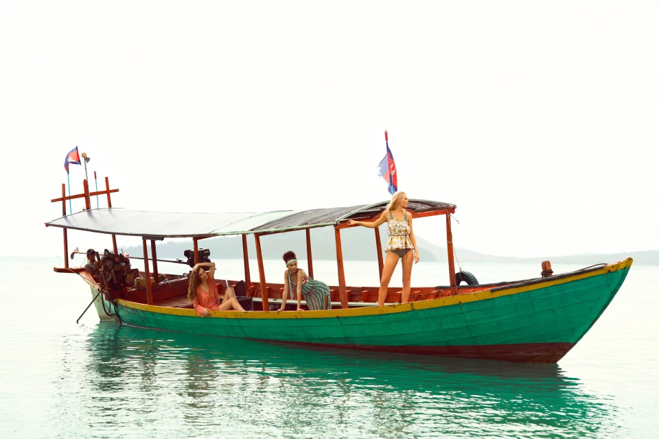 Three women on a green and brown boat in the ocean, under a bright sky, enjoying a sunny day on the water with island backdrop.