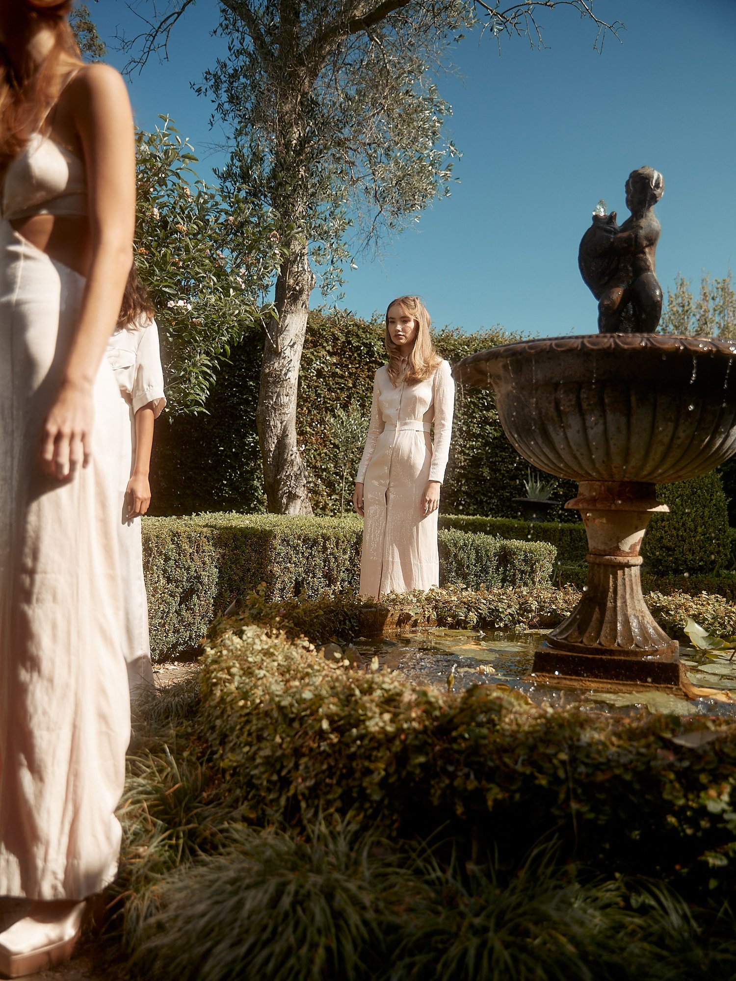 Three women in light-colored dresses pose in an outdoor garden with a fountain under a clear blue sky in soft, natural light.