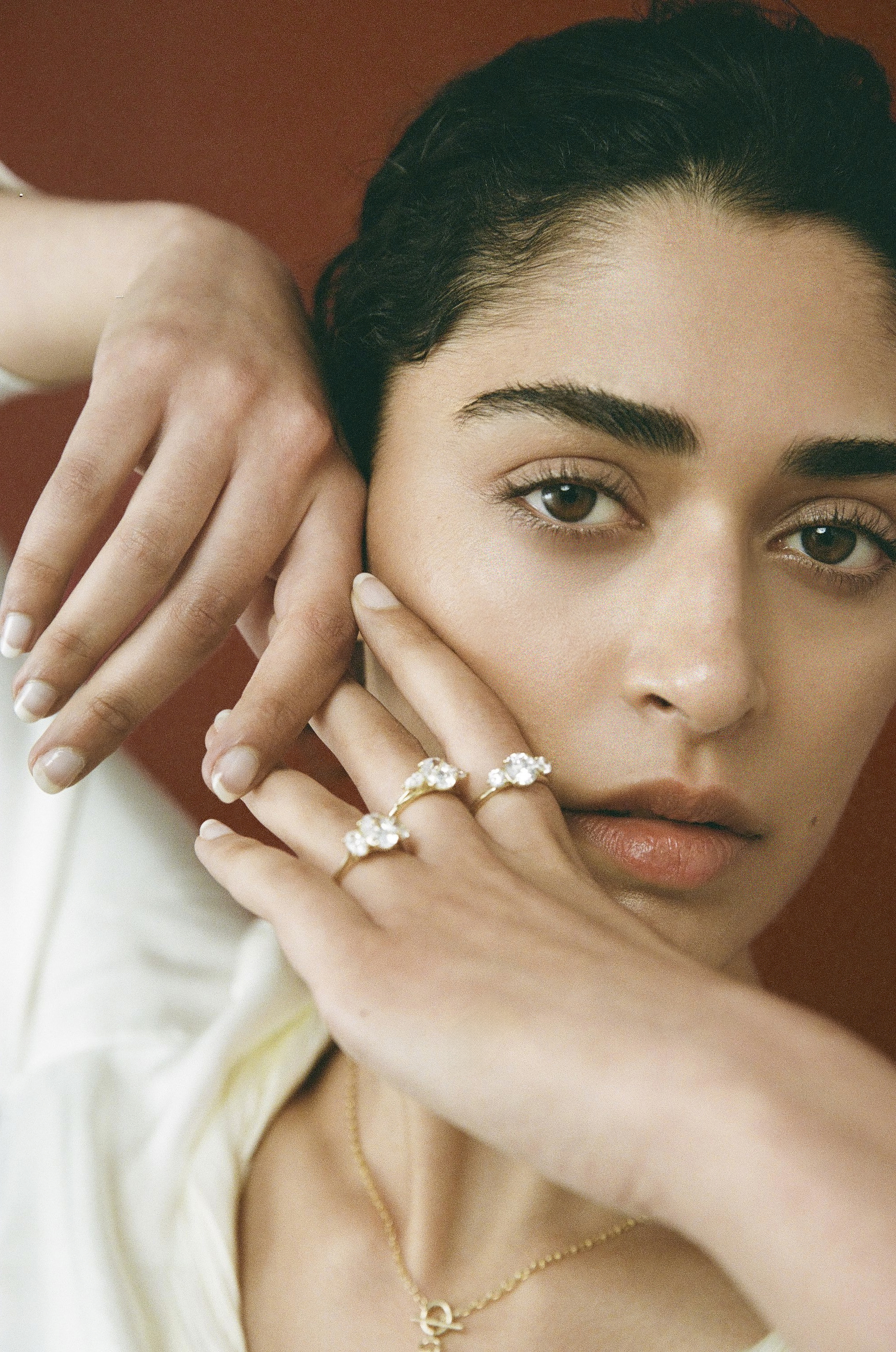 Woman with dark hair and rings poses against a red background wearing gold jewelry and a white shirt, showcasing rings on fingers.
