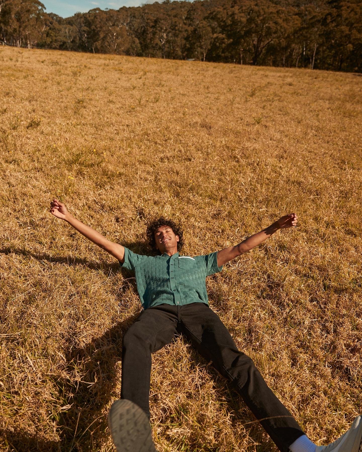 Young adult male lying in a grassy field with arms outstretched under a sunny sky, trees in the background creating a serene outdoor scene.