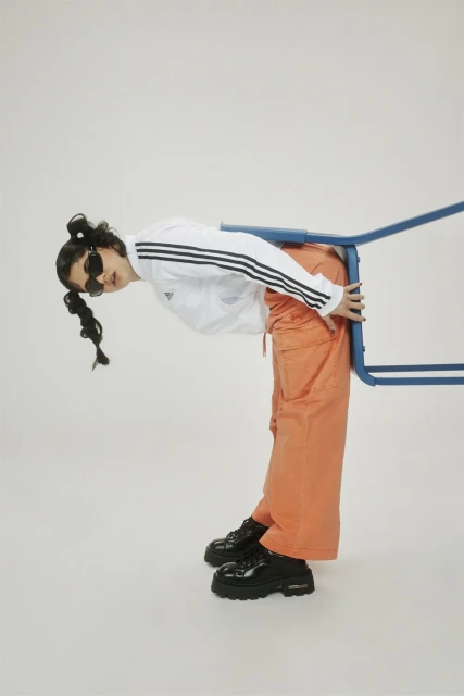 Studio shot of a woman in orange pants and an Adidas jacket bending over a blue chair against a white backdrop, wearing sunglasses.
