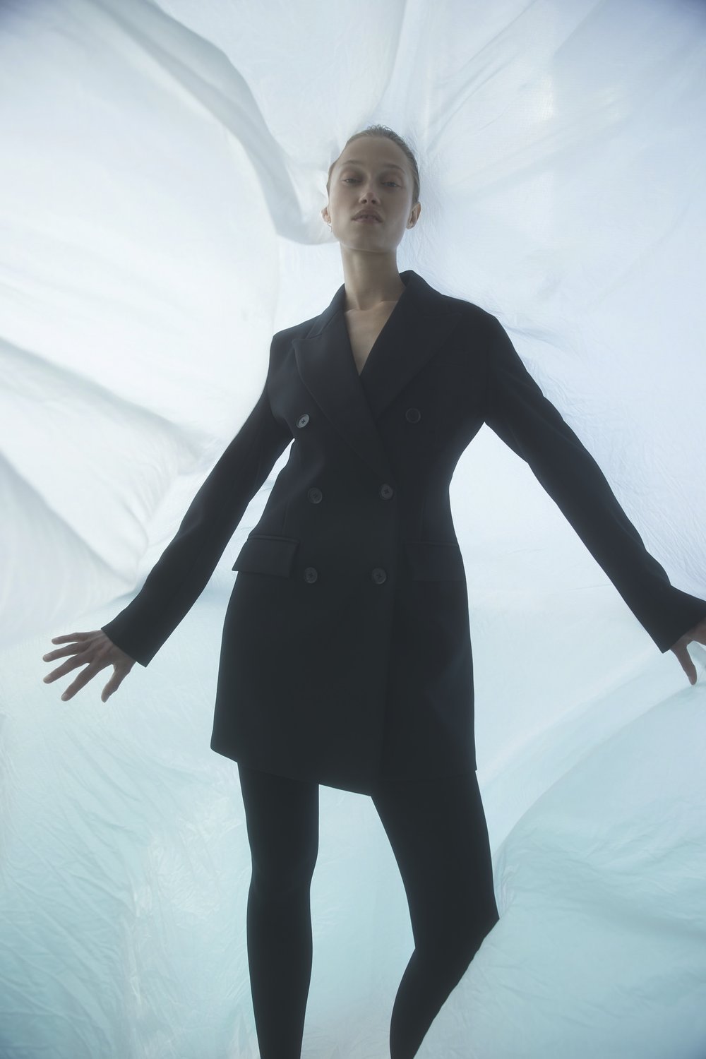 A woman in a black blazer dress and black leggings poses with her arms outstretched against a flowing white and blue backdrop.