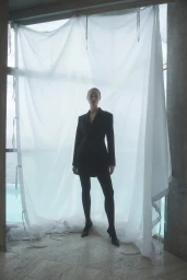 Model standing in a black double-breasted blazer and tights before a draped white fabric backdrop