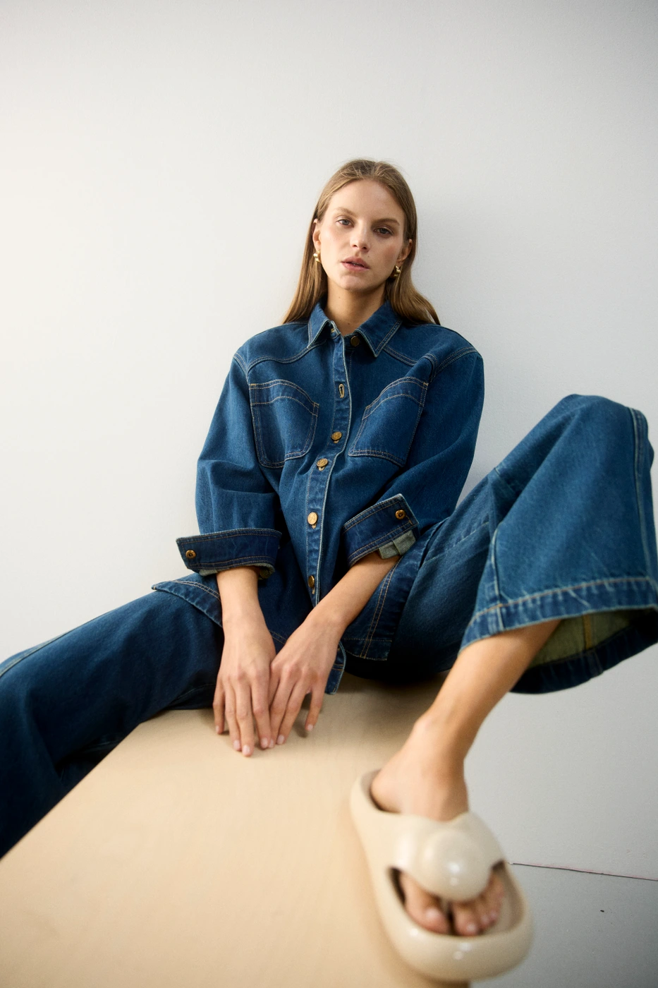 Model wearing a blue denim shirt and matching denim trousers sitting on a low wooden ledge against a white background