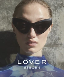 Close-up portrait of a woman wearing black oversized sunglasses and a blue patterned top against a textured stone wall.