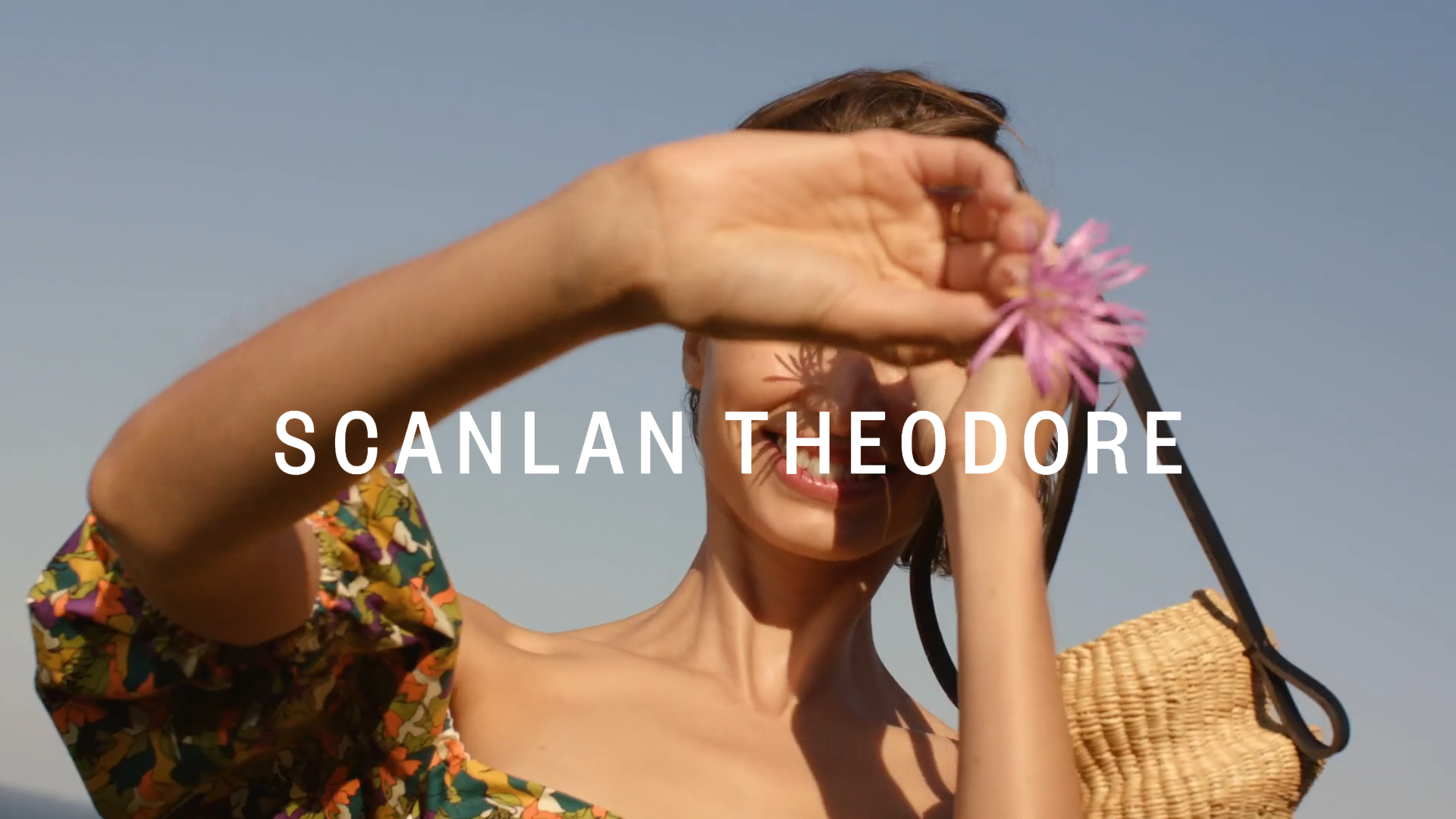 Adult woman in floral top holding purple flower outdoors against blue sky, with Scanlan Theodore text overlay.