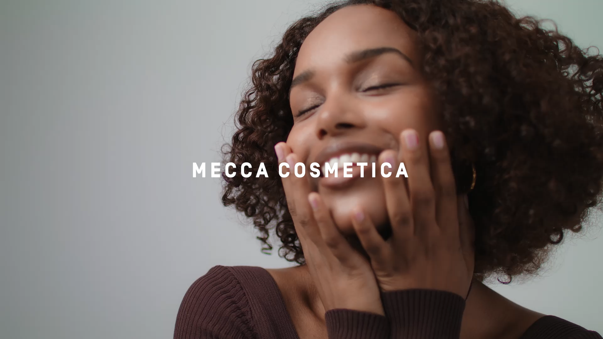 Close-up of a smiling woman with curly hair gently touching her face, set against a neutral background with the text 'MECCA COSMETICA'.
