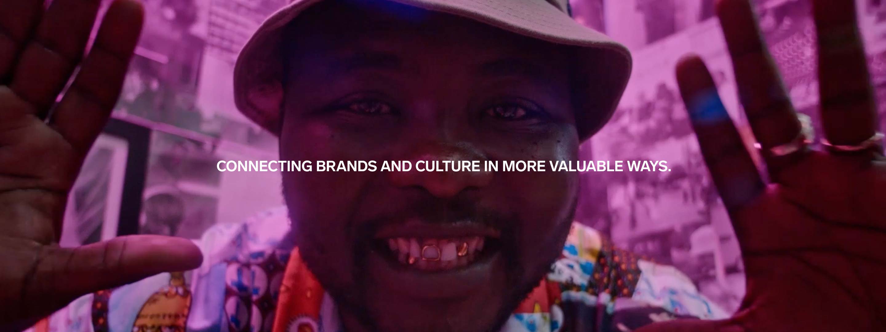 Close-up of a smiling African-American man with gold teeth, wearing a hat and printed shirt, surrounded by hands and purple light.