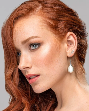 Close-up portrait of a woman with vibrant red hair, freckles, and silver teardrop earrings against a grey background.