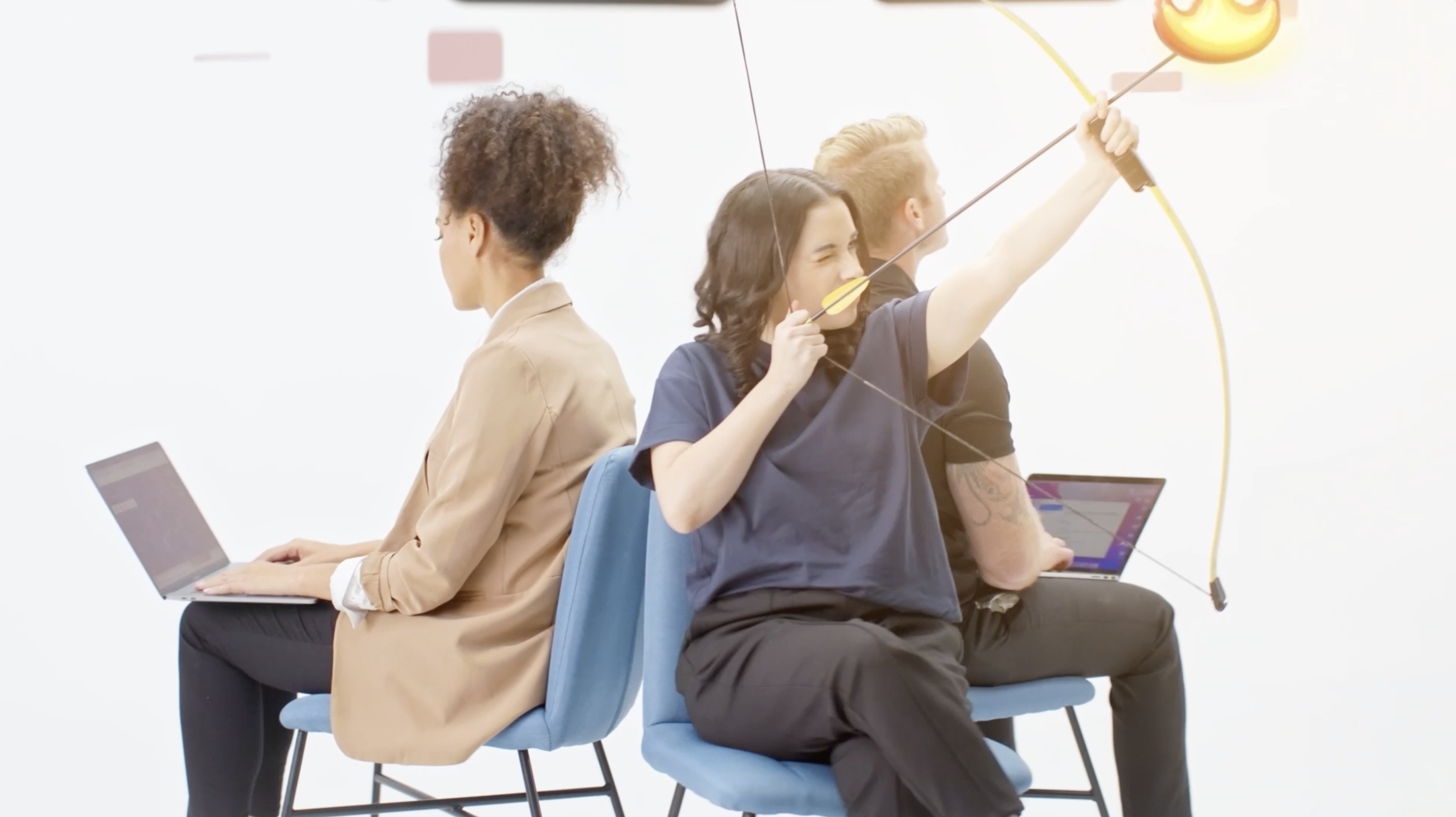 Three adults sit back-to-back on blue chairs in a bright photo studio. A woman in the center takes aim with a bow and arrow.