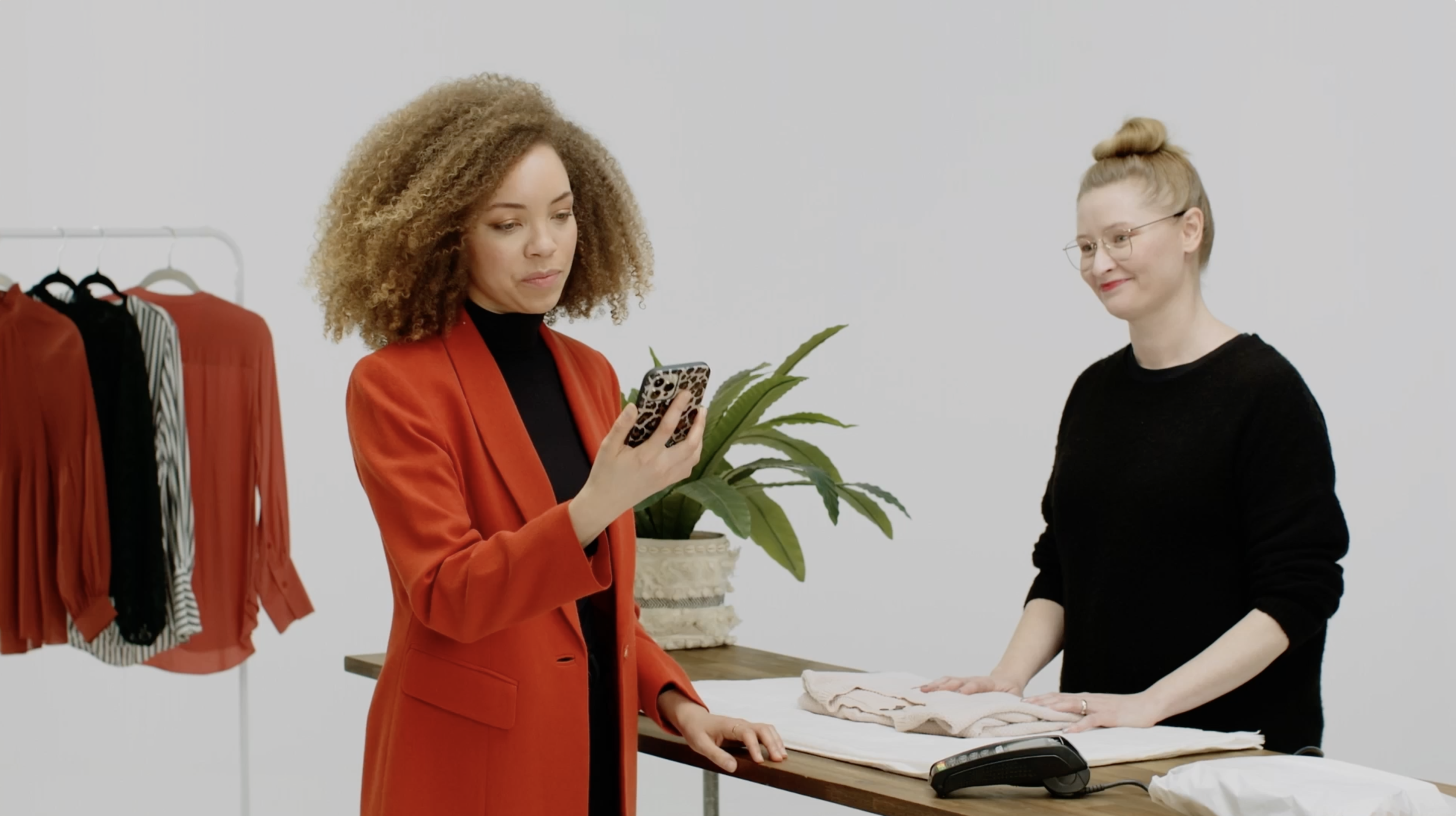 A woman in a red blazer looks at her smartphone, while another woman is folding clothes at a shop counter with potted plant.