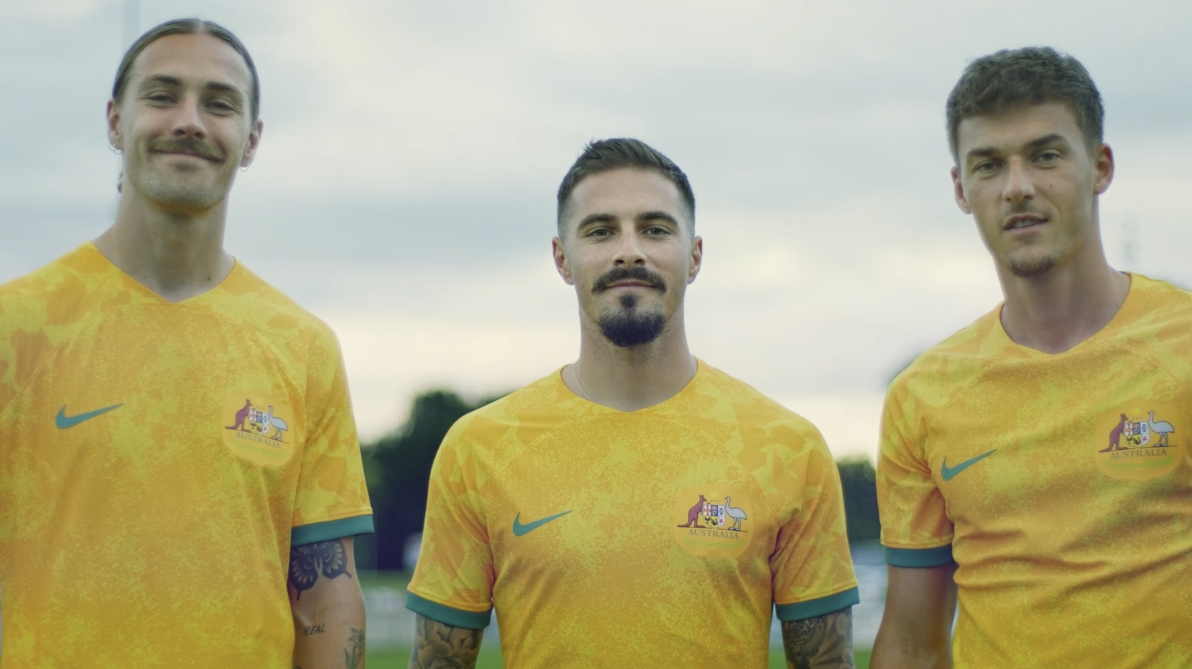 Three adult males stand outdoors in yellow Australia soccer jerseys. Close-up, eye-level angle captures their smiles against a blurred backdrop.