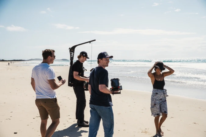 Film crew on a sandy beach, capturing a scene with a woman in a hat; sunny day, clear sky, and ocean backdrop.