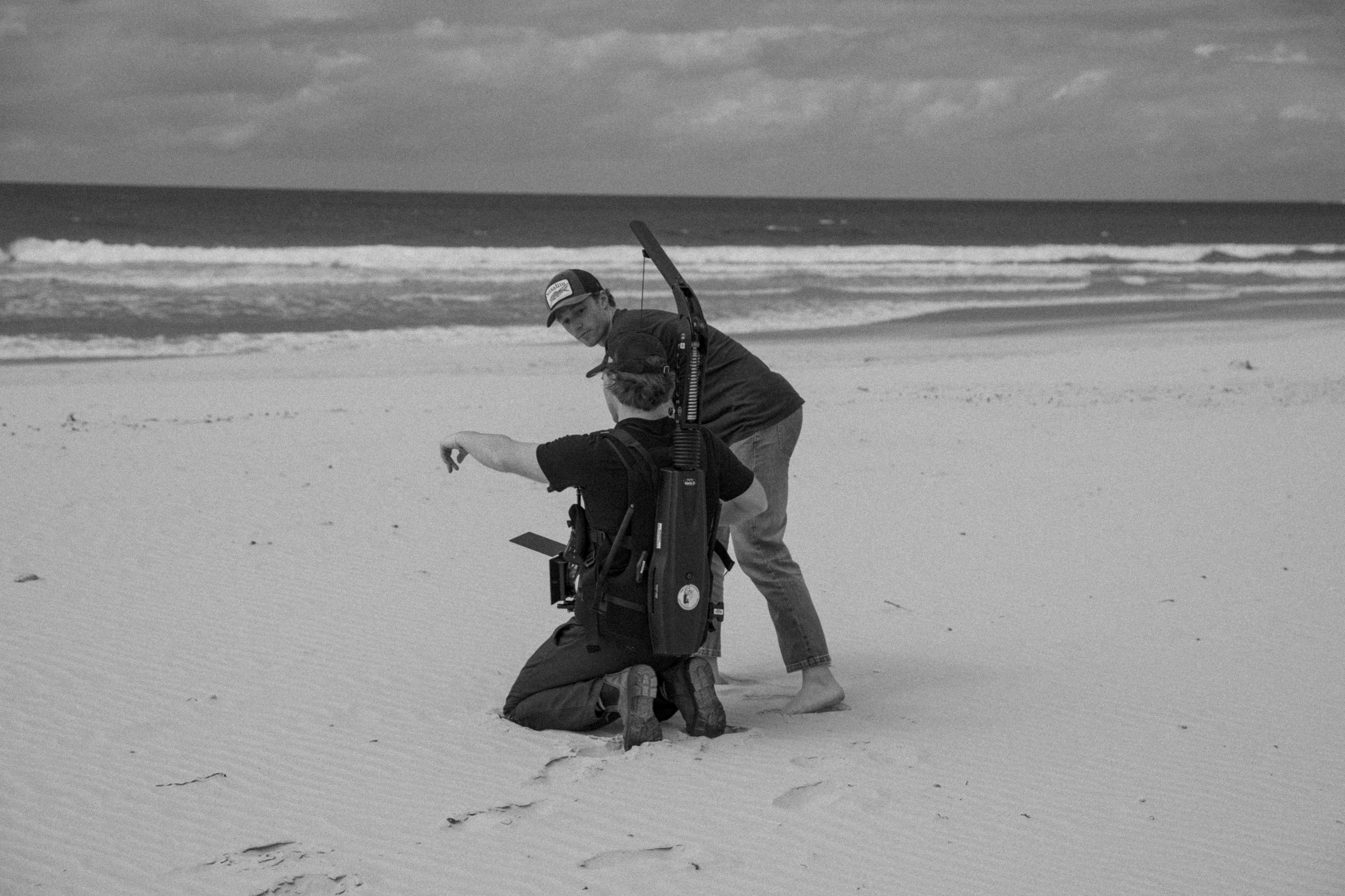 Two men on a sandy beach with camera equipment, one kneeling and pointing, the other standing and observing. Ocean and cloudy sky backdrop.