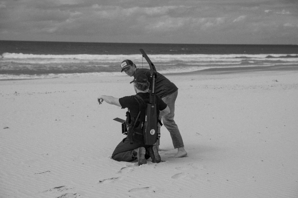 Two men on a sandy beach with camera equipment, one kneeling and pointing, the other standing and observing. Ocean and cloudy sky backdrop.