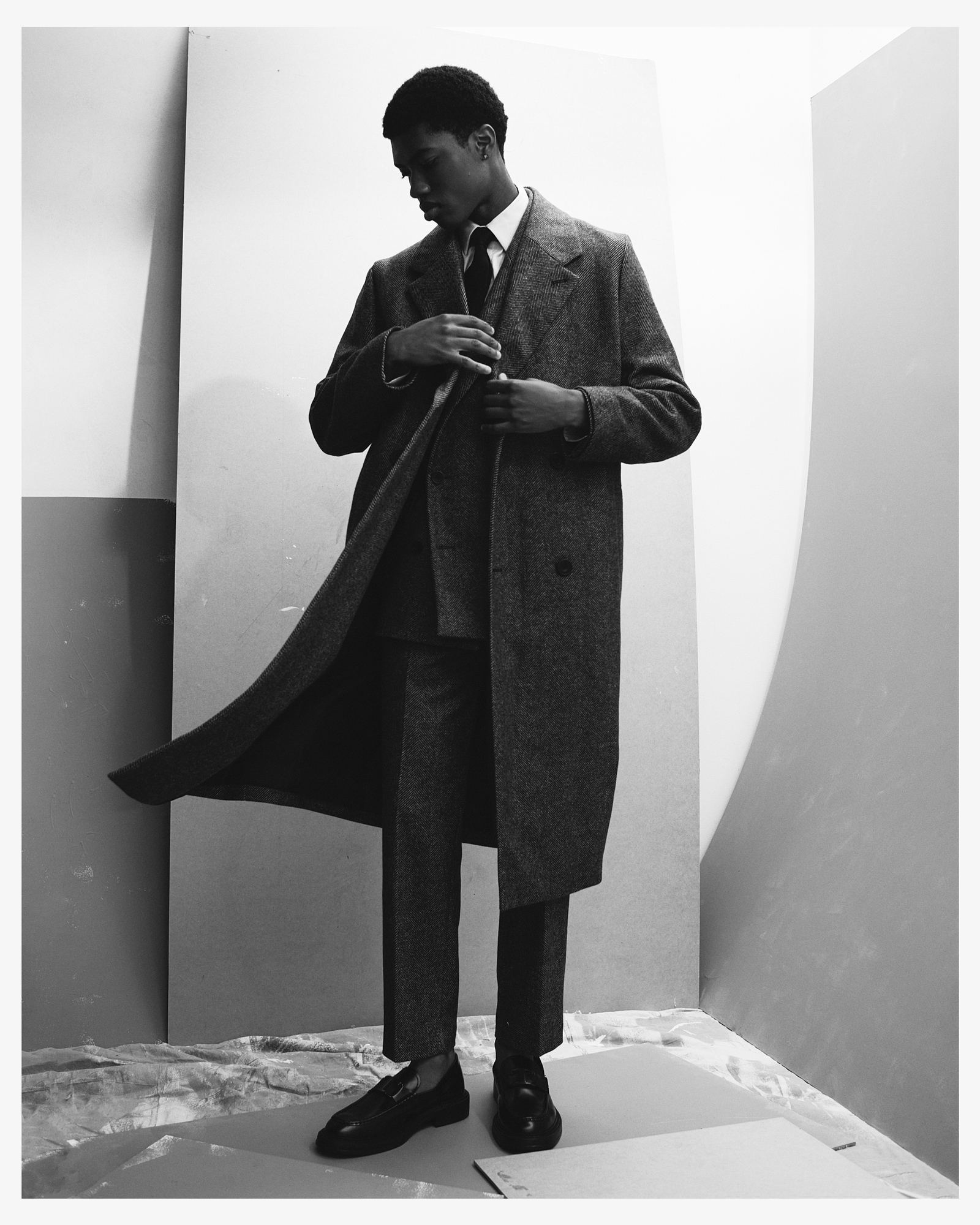 Black male model in a grey overcoat, suit, and tie strikes a pose in a studio with neutral tones, showcasing elegant fashion and style.