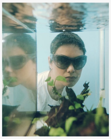 Woman with short hair wearing sunglasses looking into fish tank with plants and water reflection under bright, diffused lighting indoors.