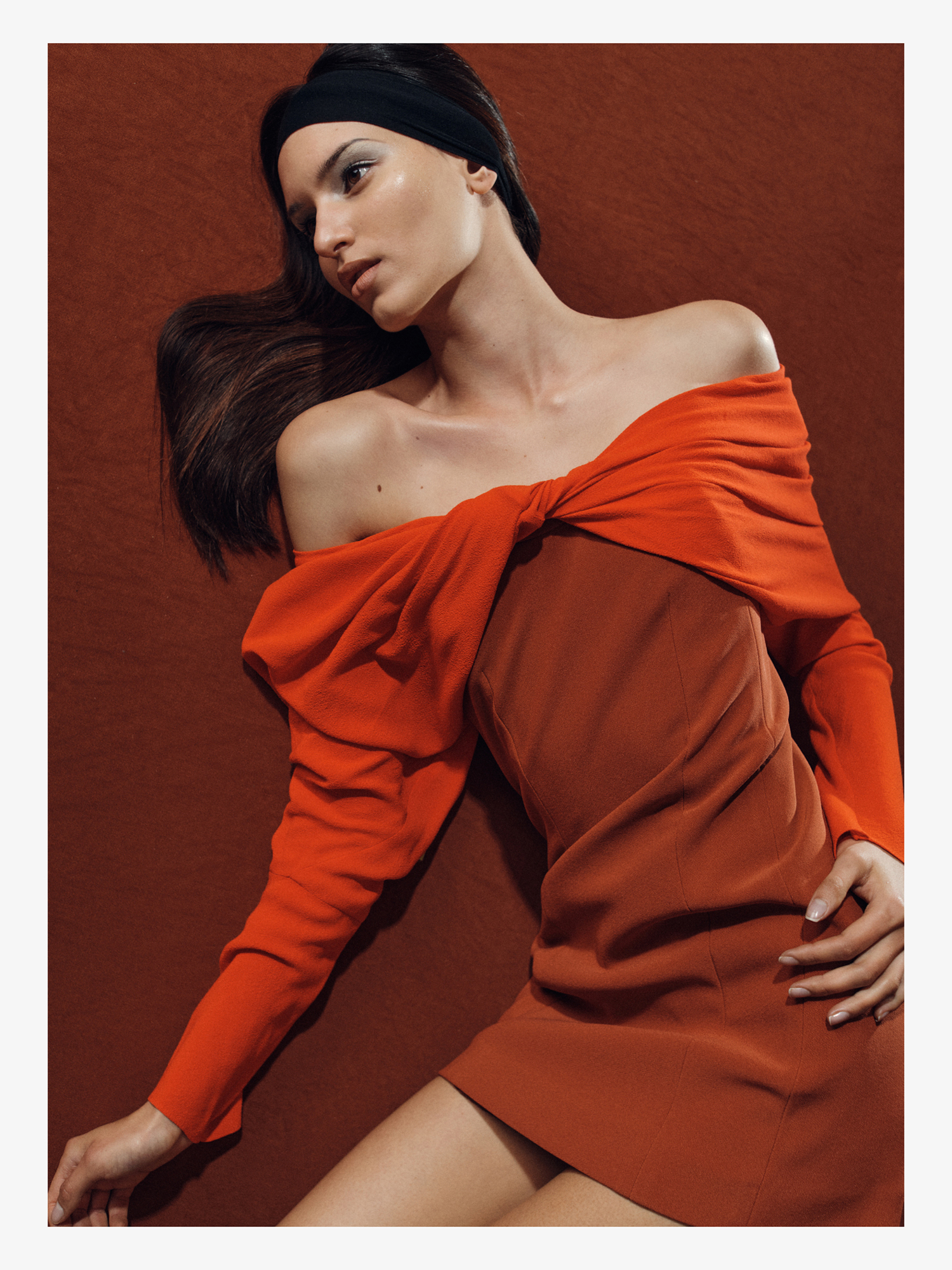 Woman in an orange off-shoulder dress lying on a textured orange background, fashion editorial style, head tilted, looking away.