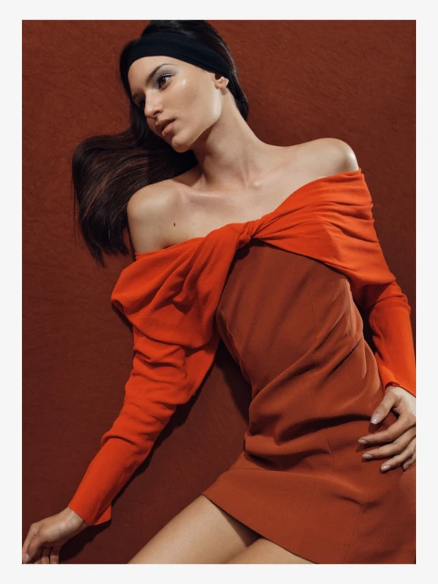 Woman in an orange off-shoulder dress lying on a textured orange background, fashion editorial style, head tilted, looking away.