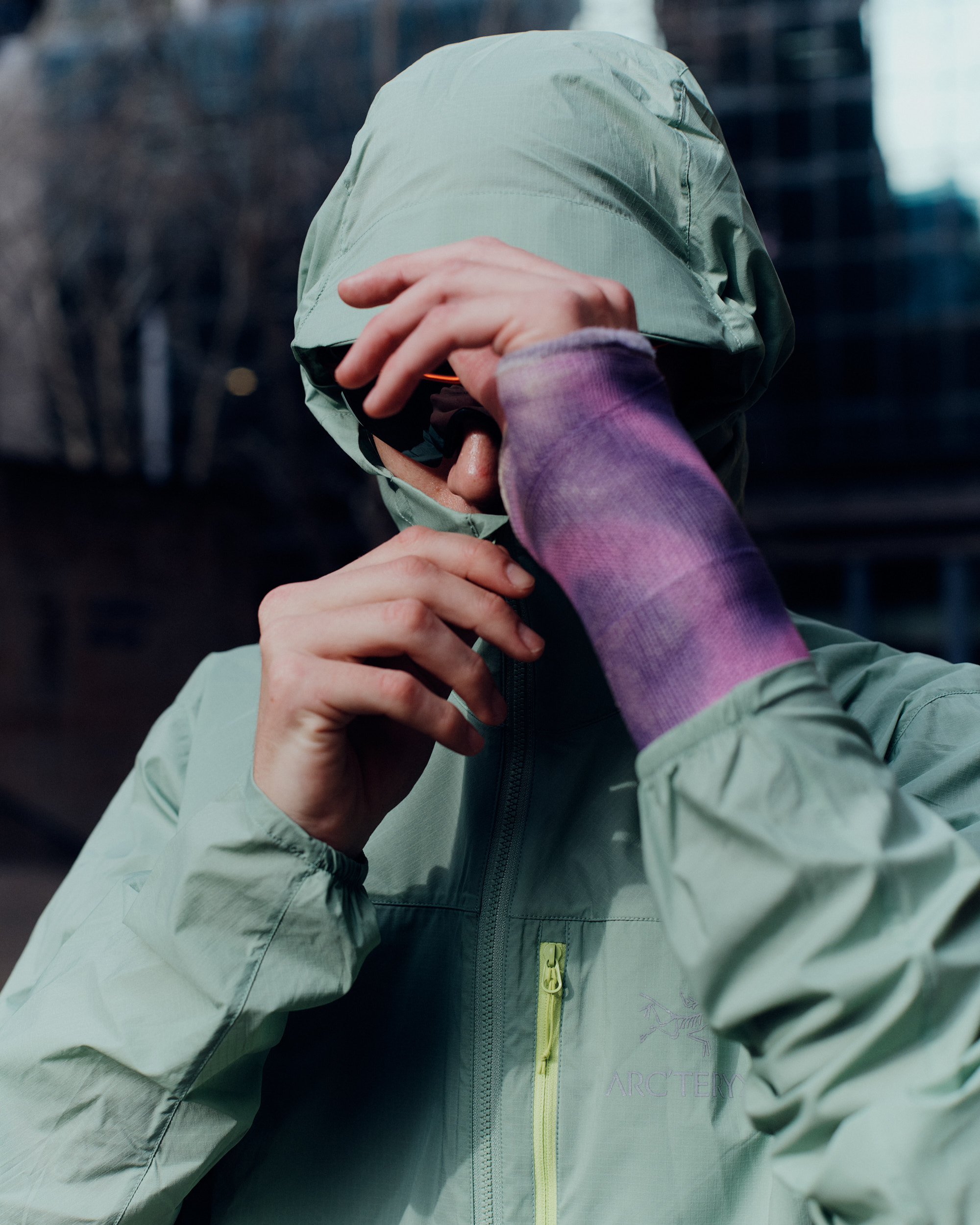 Person in mint jacket with purple cast poses outdoors against blurred building backdrop on a sunny day, displaying modern fashion.