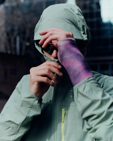 Person in mint jacket with purple cast poses outdoors against blurred building backdrop on a sunny day, displaying modern fashion.