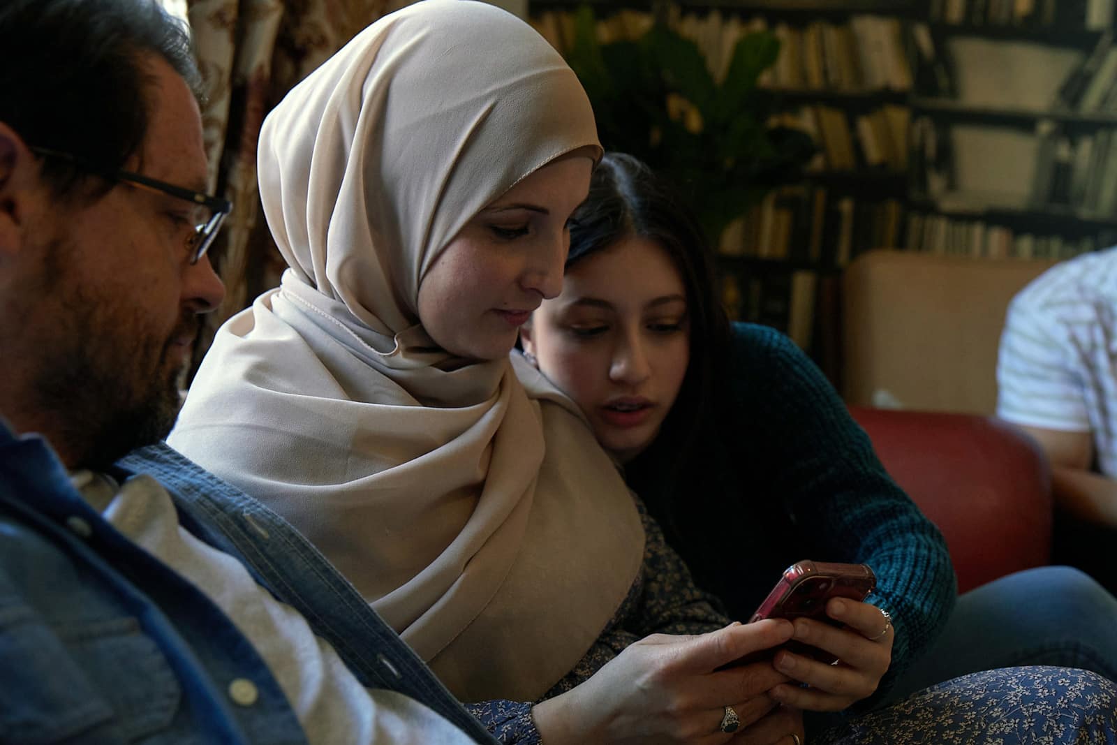 Family of three together indoors, woman in hijab holding smartphone, girl looking at screen, man with glasses nearby, casual outfits.