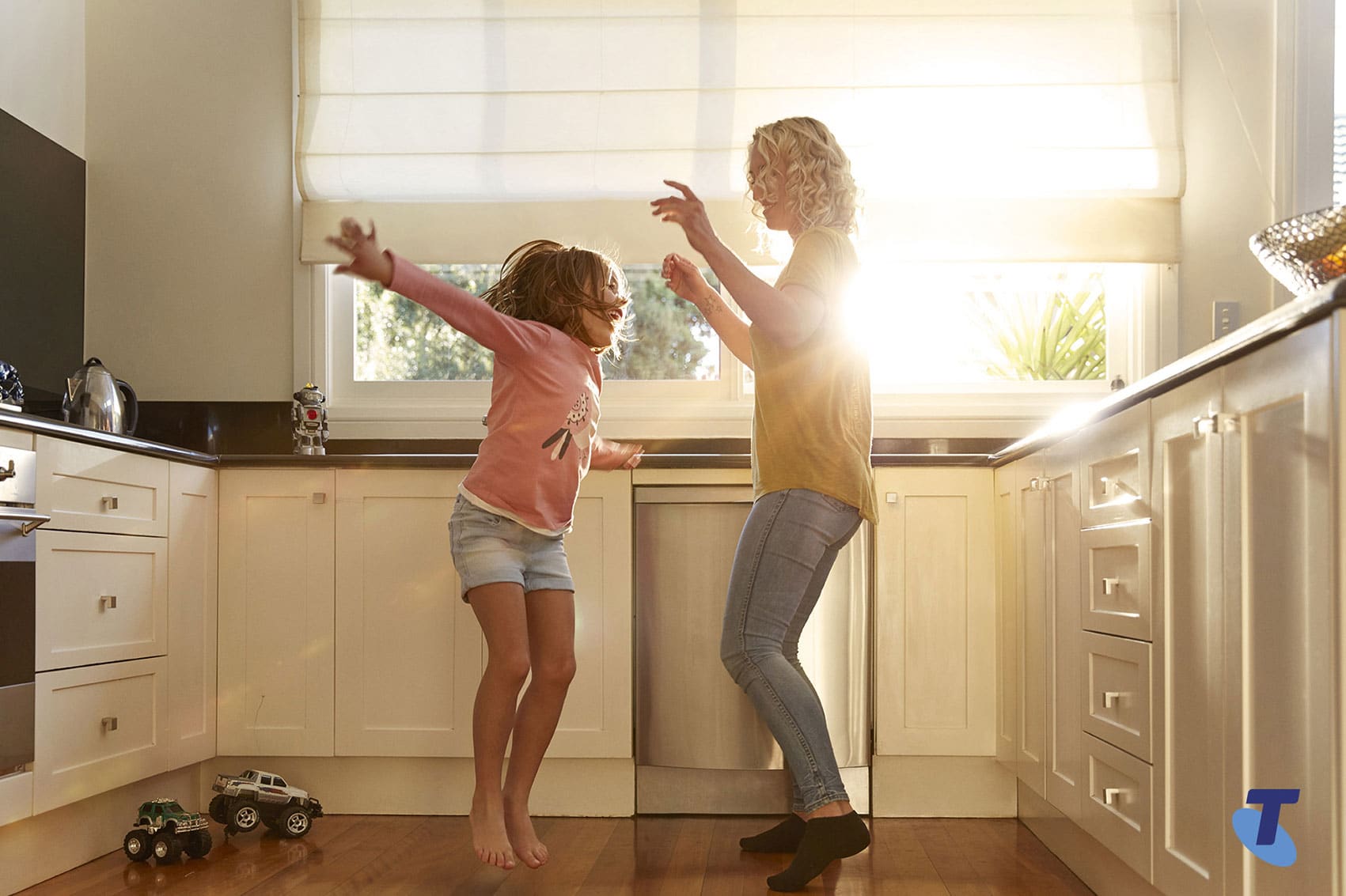 A mother and child actively play and jump in a naturally lit kitchen setting with wooden floors, white cabinets, and modern appliances.