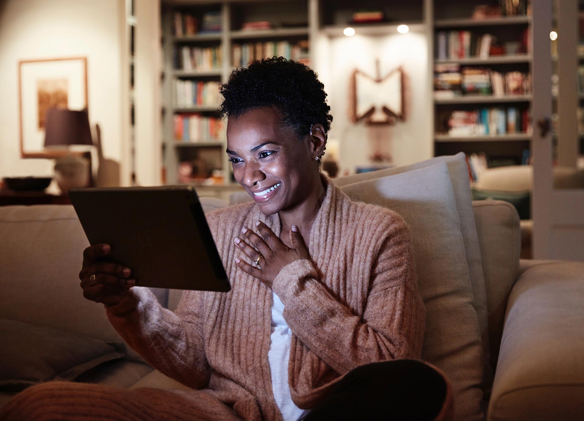Smiling woman using a tablet on a couch in a warmly lit room, with a bookshelf in the background. Casual setting, modern interior.