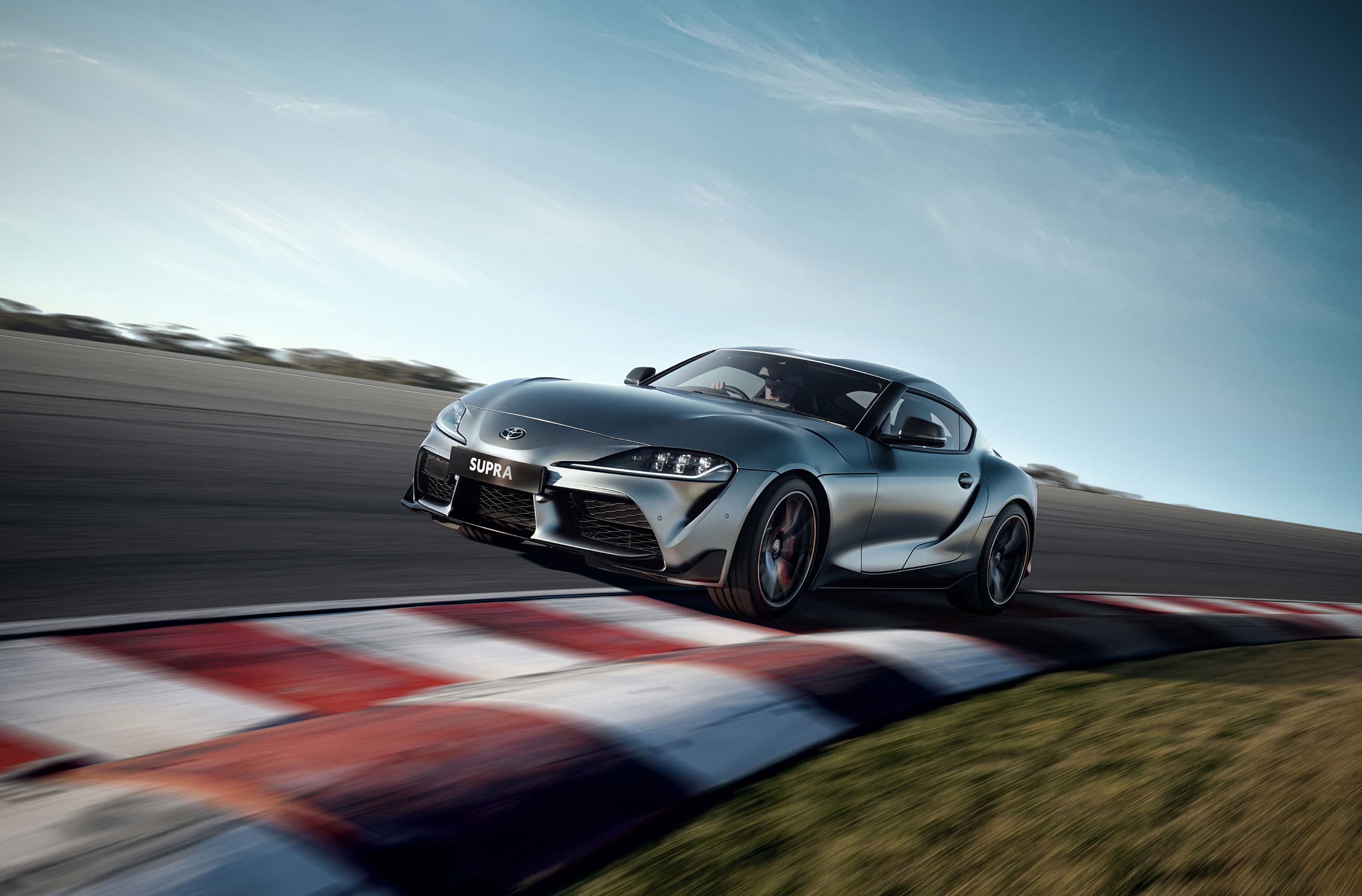 Sleek silver Toyota Supra sports car races around a track with blurred motion, showcasing automotive speed and modern design against blue sky.