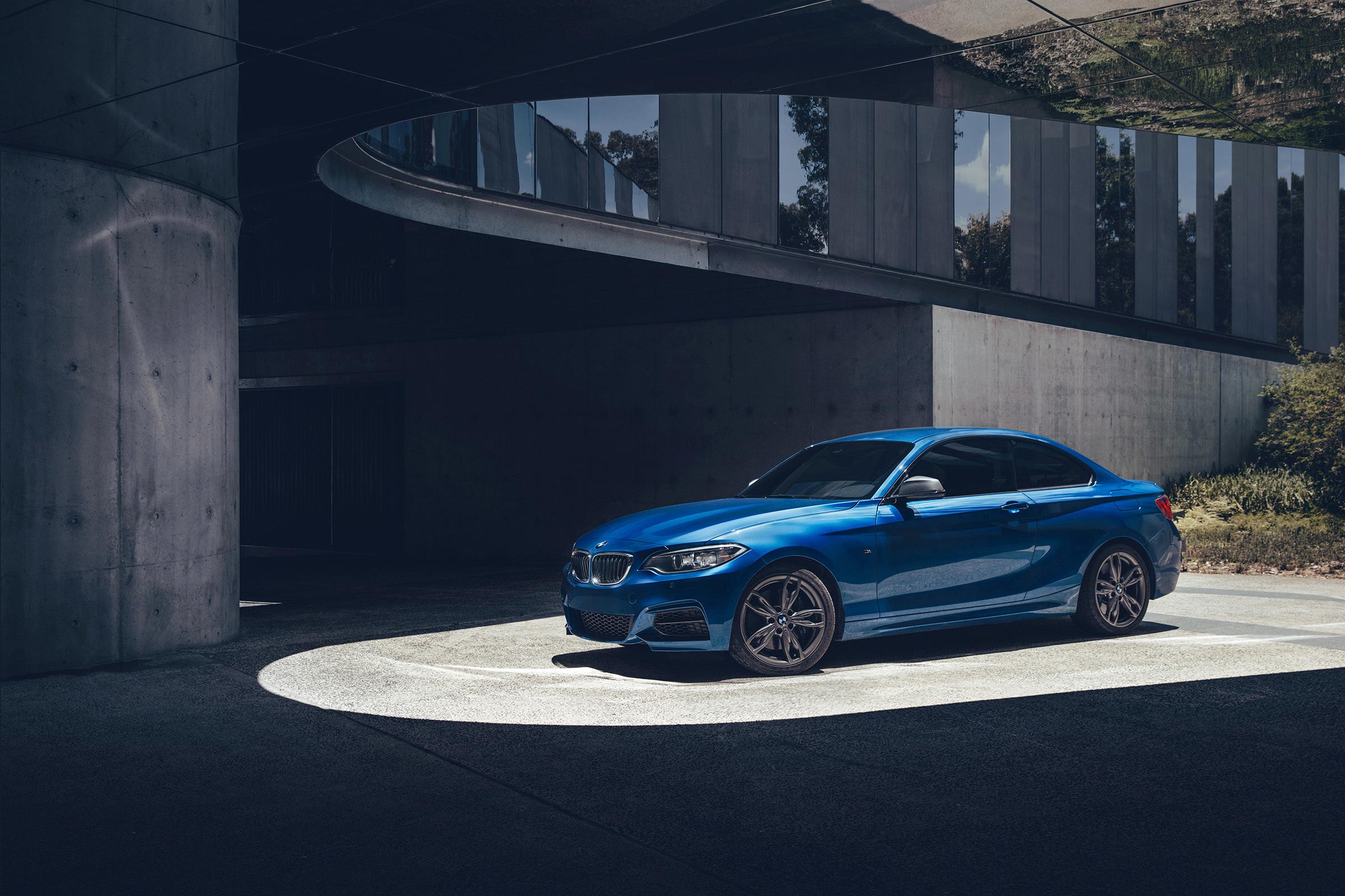 A bright blue BMW coupe is parked in an urban environment with modern architecture and dramatic lighting creating a high-contrast scene.