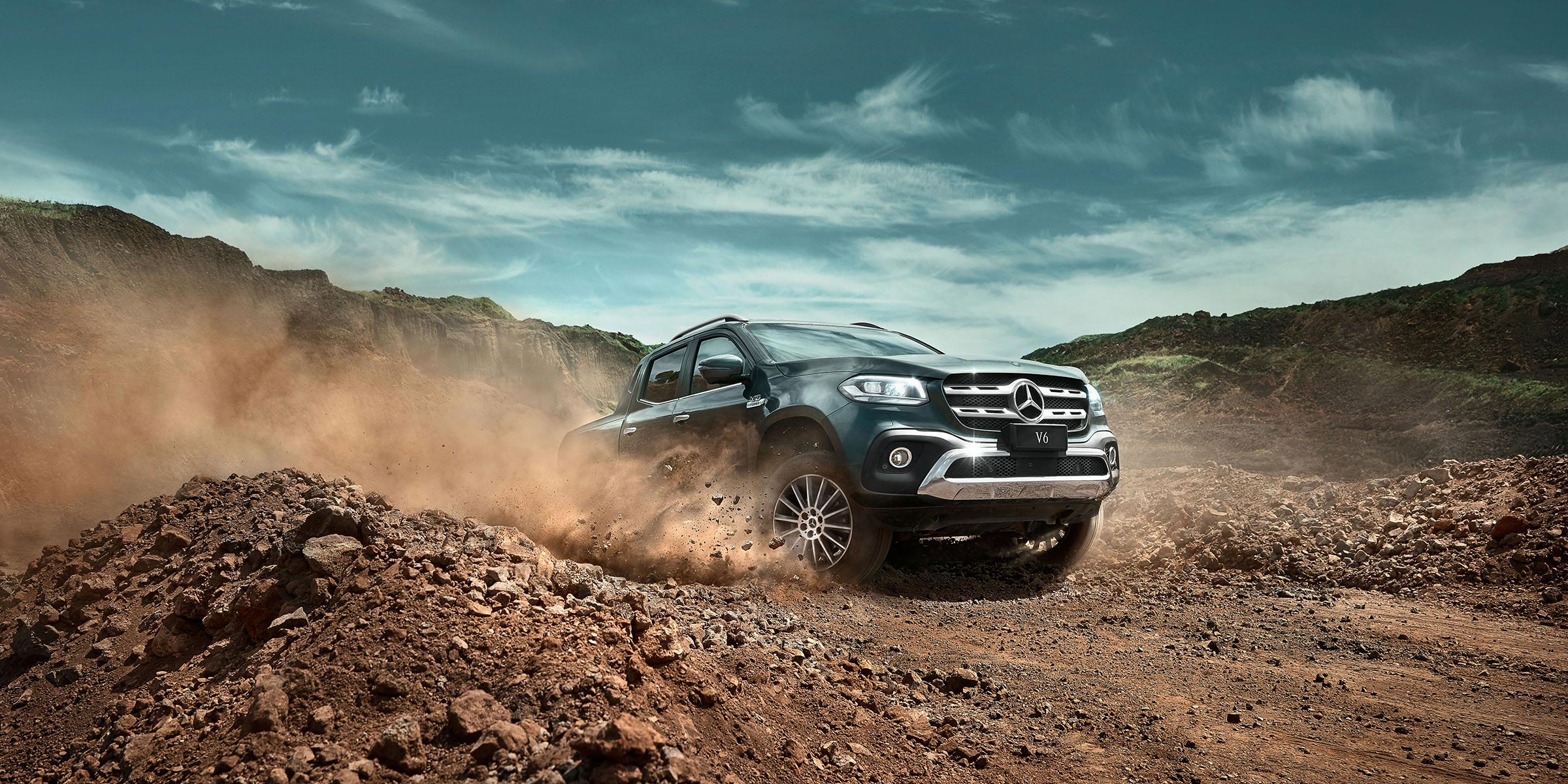 Dark grey Mercedes-Benz pickup truck driving on a dirt road with a mountain range and cloudy sky in the background.